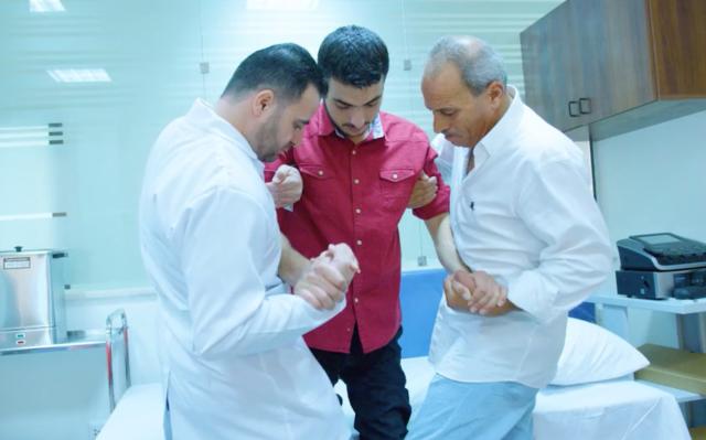 Two rehabilitation professionals standing on either side of a man holding him by the arms and help him to his feet as he tries out a new prosthetic limb.